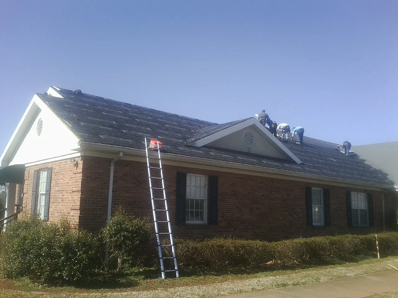 Crew tearing shingles off a brick ranch roof with a ladder set — Emergency Roof Repair work by our crew in Lake In The Hills