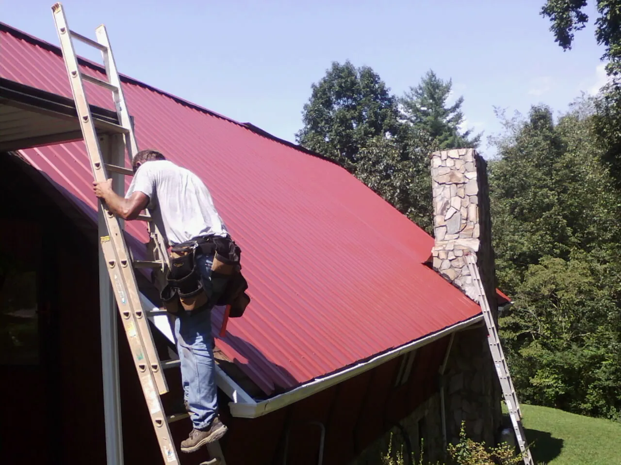 Roofer on a ladder beside a red metal roof and stone chimney — Soffit & Fascia Repair by Roofing Brothers in Lake In The Hills